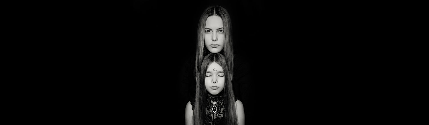 Dark aesthetic, black and white photo of a mother and her 10 year old daughter on black background. Both have long hair. The daughter has her eyes shut and a halfmoon drawn on her forehead.